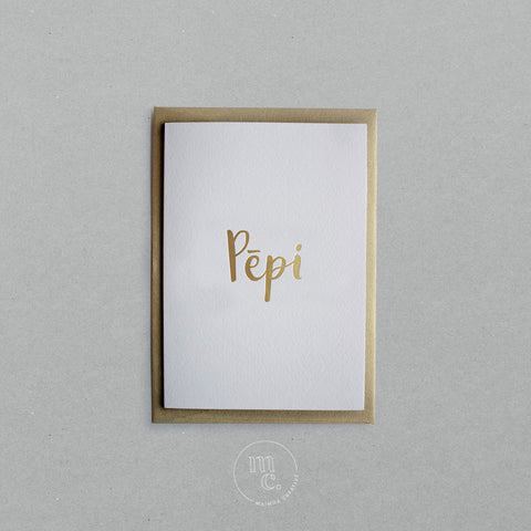 Flat lay of a premium reo Māori greeting card with the word 'Pēpi' in gold foil lettering, paired with a gold envelope