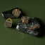 Gold pin collection with kete, tiki, kawakawa leaf and huia feather design on a dark green pounamu surface.