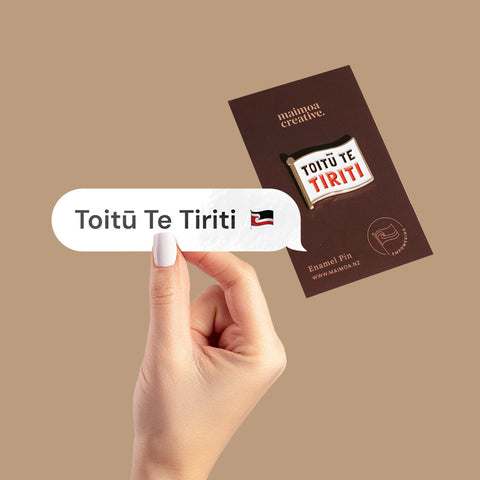 Hand holding a vinyl decal with "Toitū Te Tiriti" in a speech bubble design, with a bold enamel pin in the background with same message.