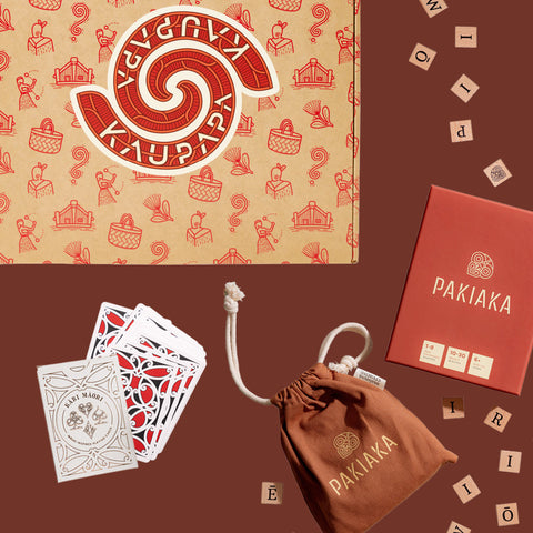 Kaupapa Game, Pakiaka word game and Kāri Māori playing cards bundle displayed with game boxes, wooden letter tiles, a cotton drawstring bag and deck of playing cards, on a brown background