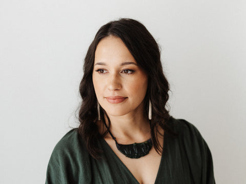 Māori woman wearing a taonga, whalebone earrings and dark green dress looking to the side with slight smile, headshot of Aroha Tamihana.