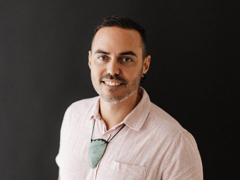 Māori man wearing a taonga and neutral shirt standing against a dark background and smiling into the camera. Head shot of Hamuera Tamihana.