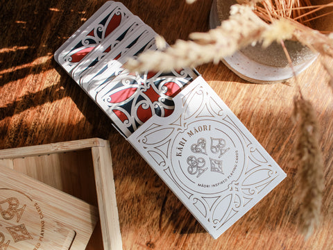 Māori-inspired playing cards displayed on a wooden table, partially fanned out next to a ceramic vase and dried grass in sunlight