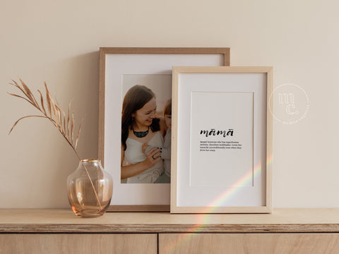 Two framed prints on a wooden shelf; on the left, a mother-daughter photo, and on the right, a 'māmā' definition print, with a vase and dried flowers beside them.