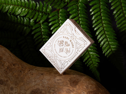 Kāri Māori playing cards balancing on a natural hue maori gourd, set against lush ferns in a moody, dark setting.