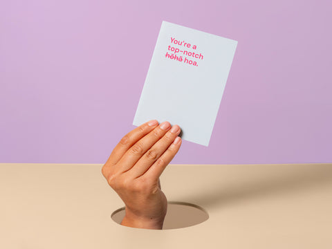 Hand poking through a hole holding a greeting card with the text 'You're a top-notch hōhā-hoa' against a pastel purple and beige background.