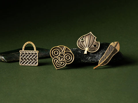 Four gold Māori pins with different designs on dark pounamu on a green background